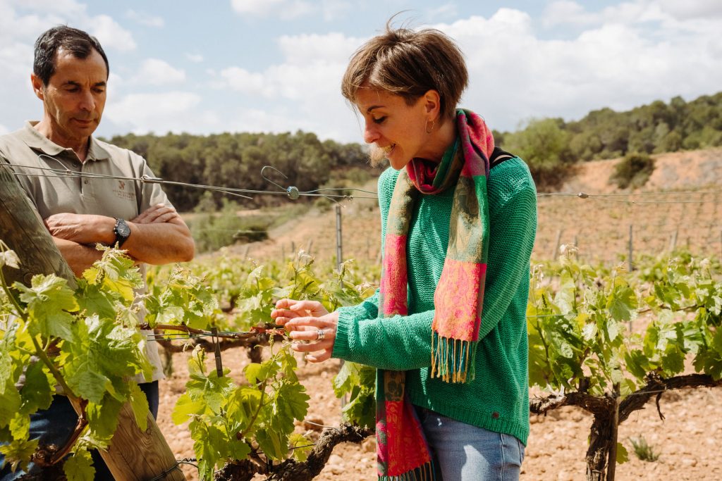 A woman in a green sweater and scarf inspects grapevines in a Montserrat vineyard, smiling, with a man observing nearby.