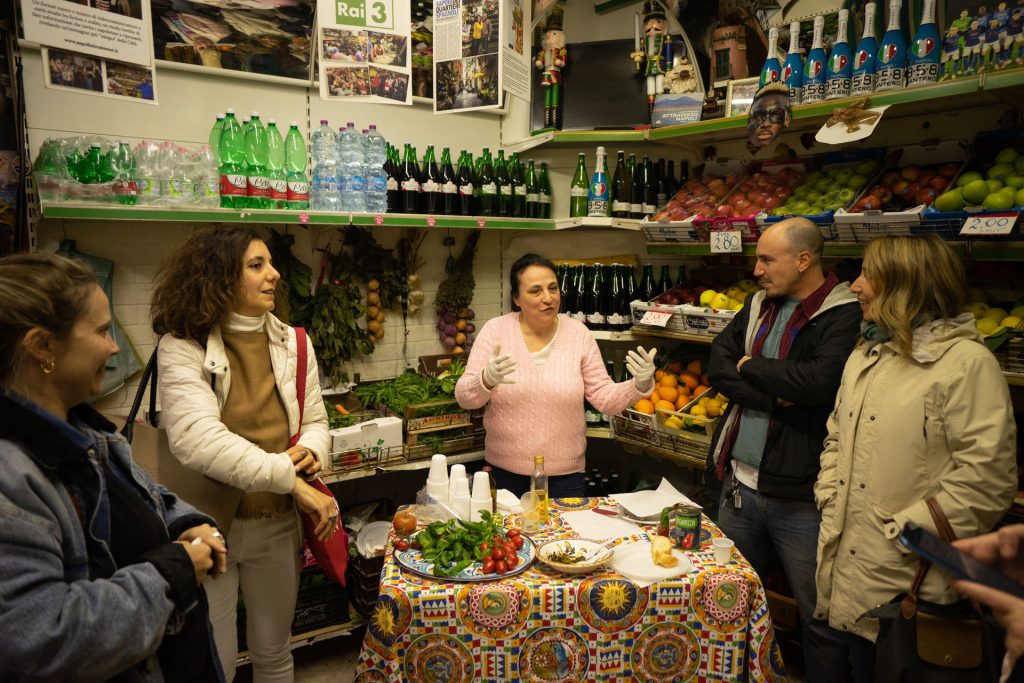 A woman in a pink sweater and gloves talks with four people around a decorated table at a vibrant Naples produce shop.