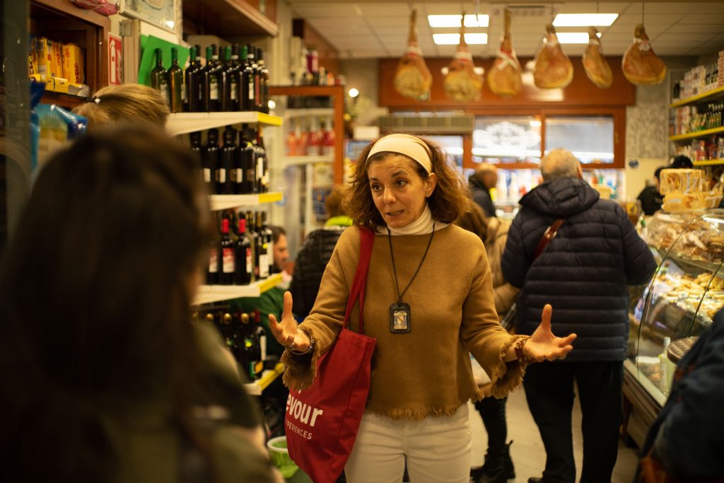 A woman with a red bag gestures while chatting with friends inside a bustling deli in Naples, surrounded by meats and bottled goods.