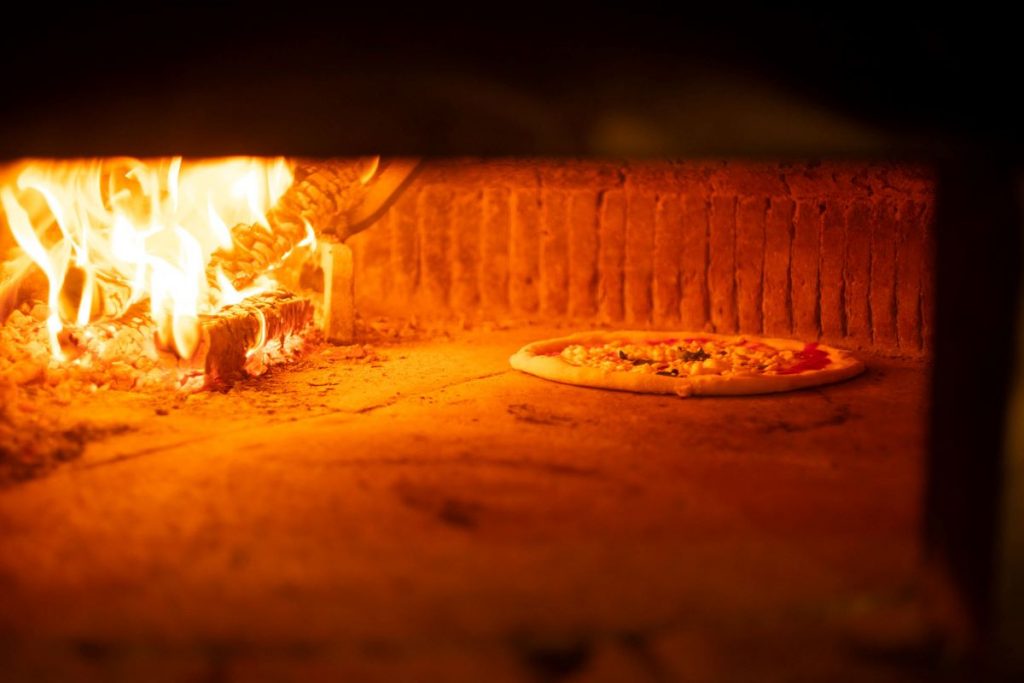 A Neapolitan pizza bakes on a stone floor beside bright flames and logs in a traditional wood-fired oven in Naples.