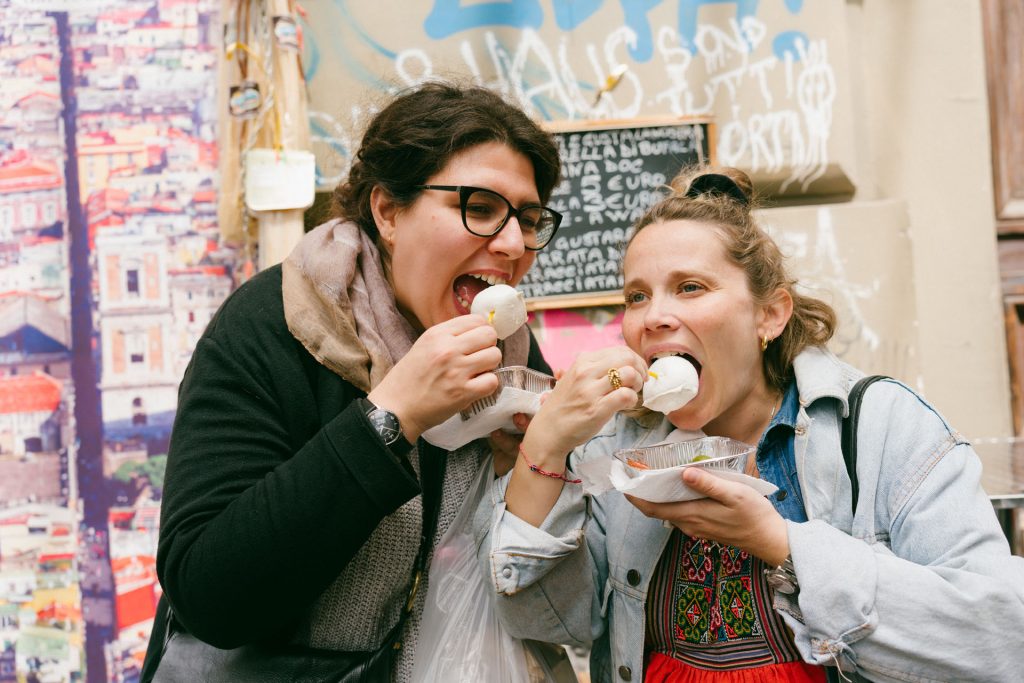 Two women smile and eat frozen treats with spoons beside a vibrant graffiti wall in Naples, celebrating after a pizza quest.