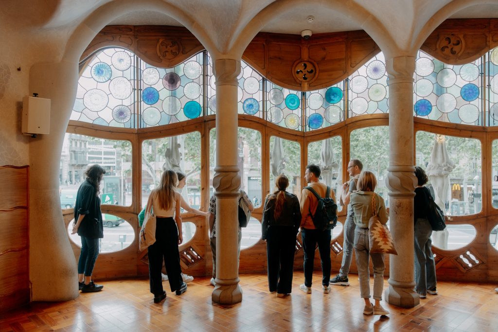 Several people stand in Casa Batlló, Barcelona, admiring the city view through Gaudí’s colorful stained glass and curved wooden windows.