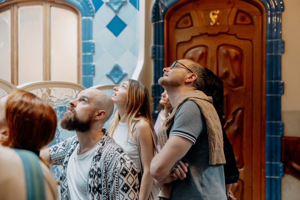 A group of people in a grand, historic hall with blue and wood accents gaze upward, possibly admiring the intricate ceiling.