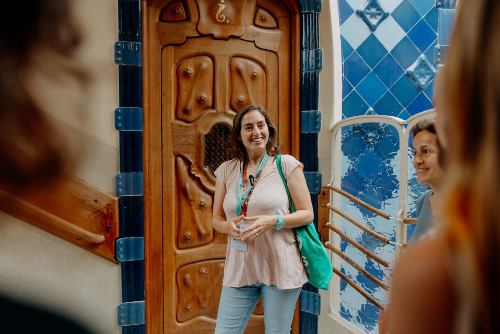 A woman carrying a green bag smiles and talks to a group near an ornate wooden door and blue-and-white tiled wall, possibly in Lisbon.