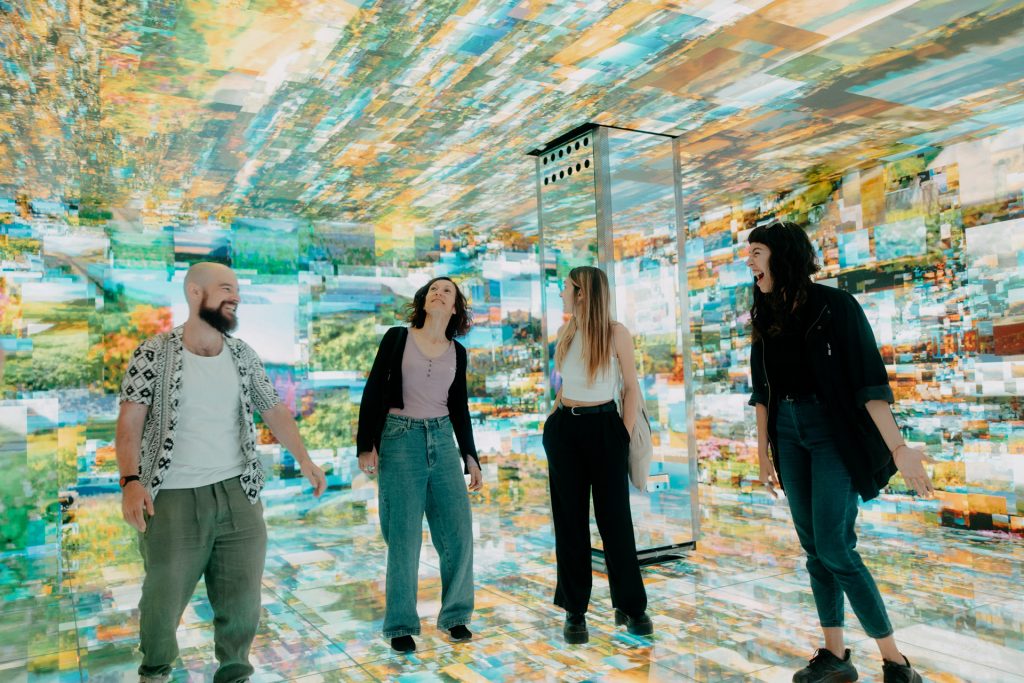 Four people smile and laugh together in a brightly lit room, surrounded by colorful, floor-to-ceiling digital projections at teamLab Borderless Tokyo.