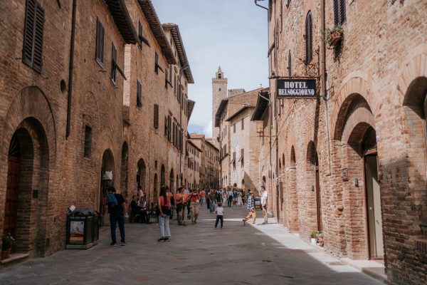 Pedestrians stroll along Via San Matteo in San Gimignano, Italy, passing Hotel Bel Soggiorno’s sign and medieval tower.