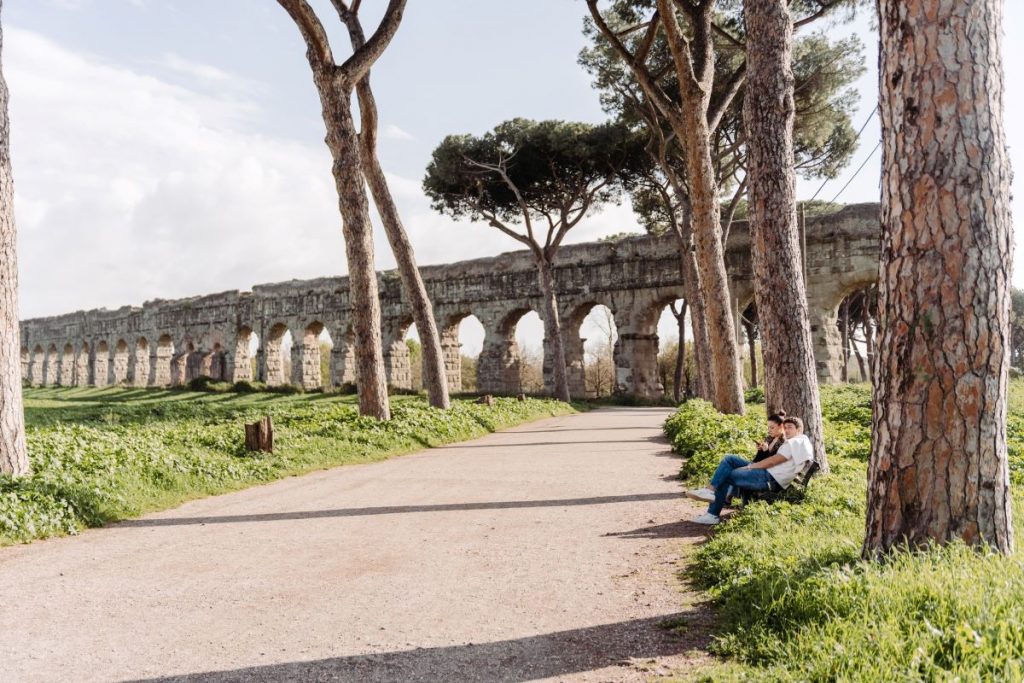 Two people rest on a bench beneath tall pines near Rome’s ancient stone aqueduct, surrounded by lush green grass.