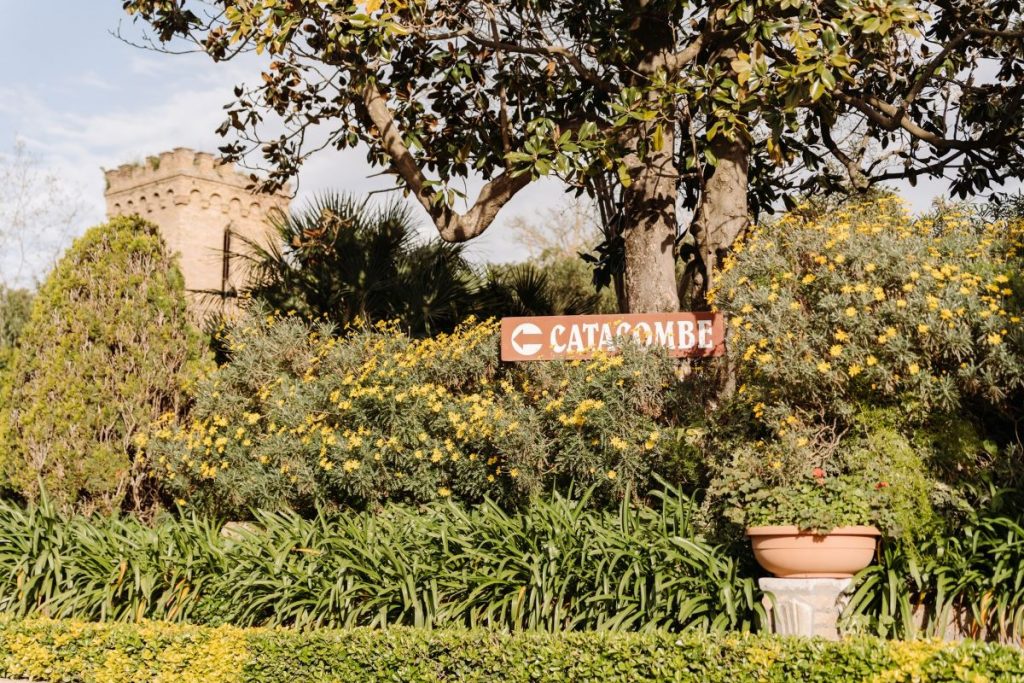 A brown sign labeled Catacombe points right, surrounded by green plants and yellow flowers near a partially hidden Rome stone tower.