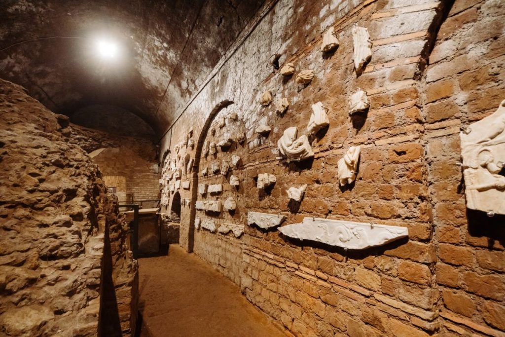 A dimly lit underground corridor in Hidden Rome features ancient carved stone reliefs and fragments mounted on brick walls.