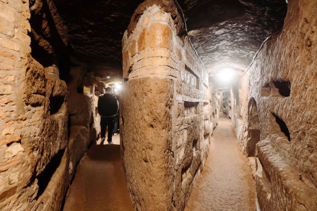 Two people walk through a narrow, dimly lit stone tunnel lined with niches, exploring Romes ancient catacombs beneath the city.