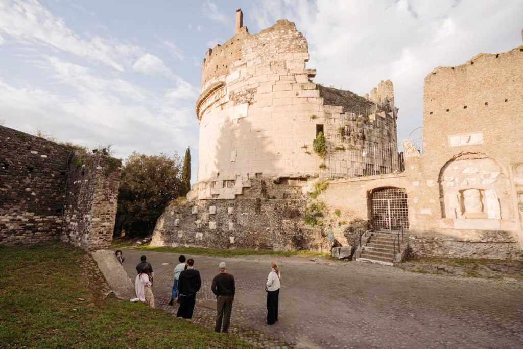 Six people explore ancient rounded stone ruins in Rome, examining weathered architecture together under a partly cloudy sky.