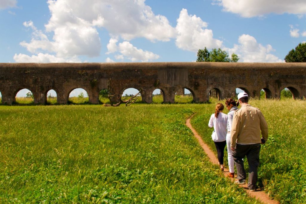 Three people stroll along a dirt path toward an ancient stone aqueduct with multiple arches in the Roman countryside under scattered clouds.