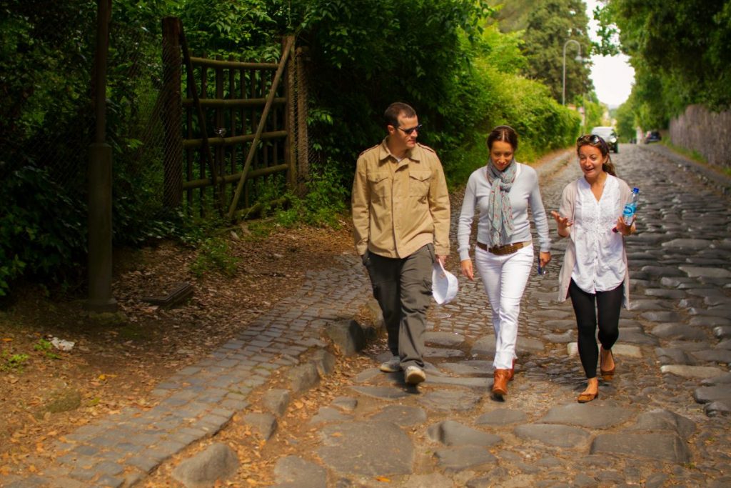 Three friends stroll along a cobblestone path in Rome, chatting and smiling, surrounded by lush greenery on a sunny afternoon.