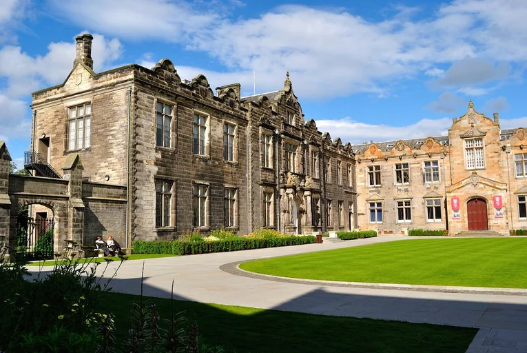 Two people relax on a bench beneath historic stone university buildings on Edinburgh’s campus, enjoying the peaceful, sunny green lawn.