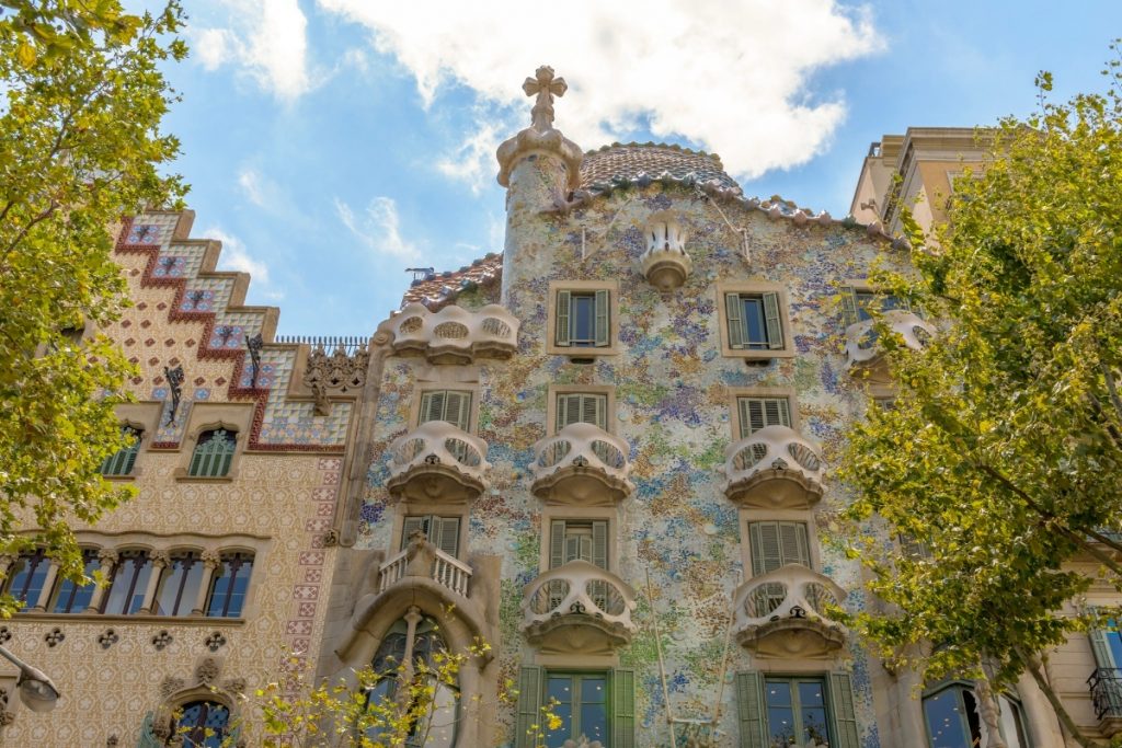 Casa Batlló in Barcelona displays Gaudí’s whimsical design, with vibrant mosaic tiles, wavy balconies, and sunlight filtering through surrounding trees.