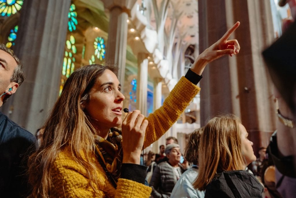 A woman in a yellow sweater leads a guided tour, gesturing upward inside a grand hall with stained glass windows.