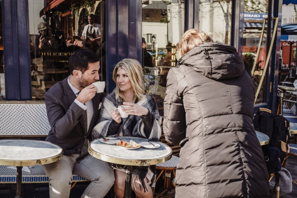 A couple enjoys coffee and pastries at a Paris café while a person in a dark coat photographs the bustling street nearby.