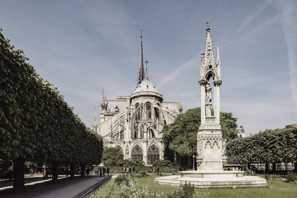Notre-Dame Cathedral in Paris stands behind manicured trees and a garden, with a tall stone fountain under clear blue skies.