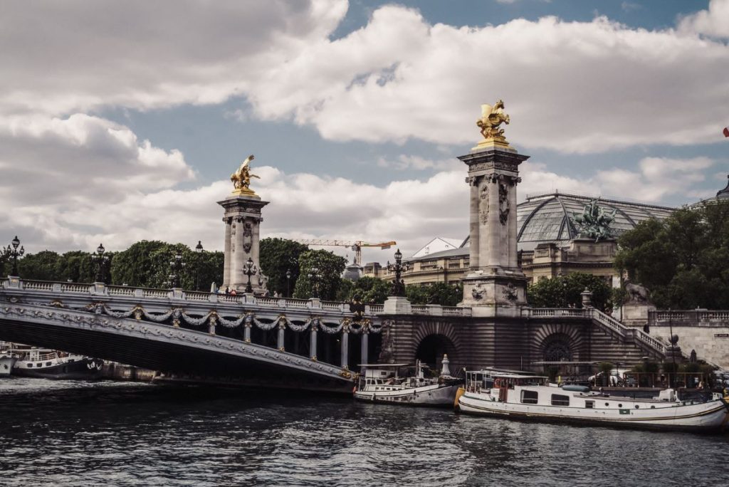 Pont Alexandre III bridge in Paris spans the Seine, adorned with golden statues, boats docked below, and historic buildings nearby.