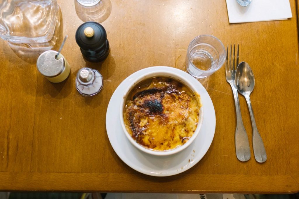 A caramelized crème brûlée rests on a white plate beside utensils and condiments, set on a wooden Paris café table.