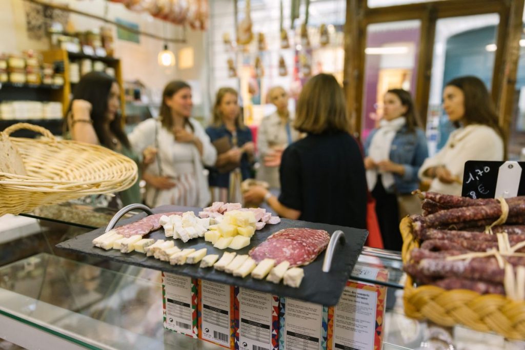 Several people chat beside a deli counter with assorted cheeses and cured meats; shelves and hanging meats create a lively Parisian atmosphere.