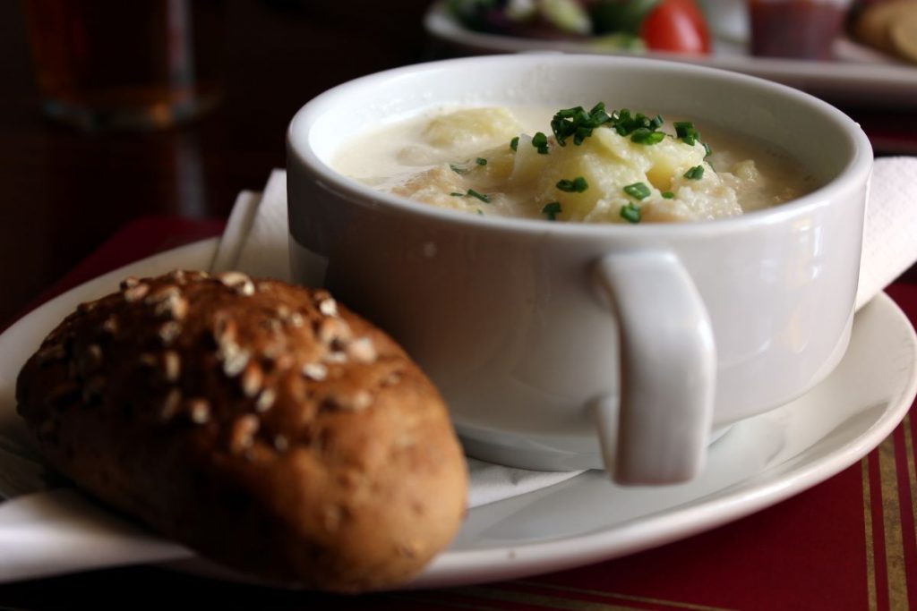 A steaming bowl of potato soup garnished with chives sits beside a seeded roll, evoking Edinburgh’s classic comfort cuisine.