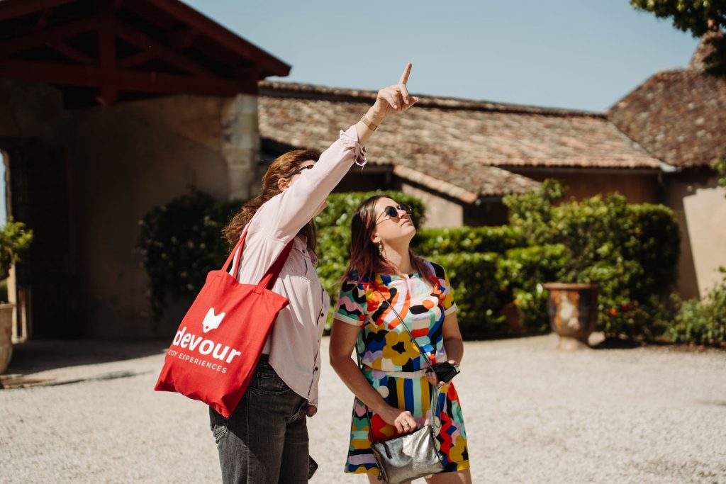 Two women stand in Bordeaux; one points upward with a red devour tote bag, both gazing at something above.
