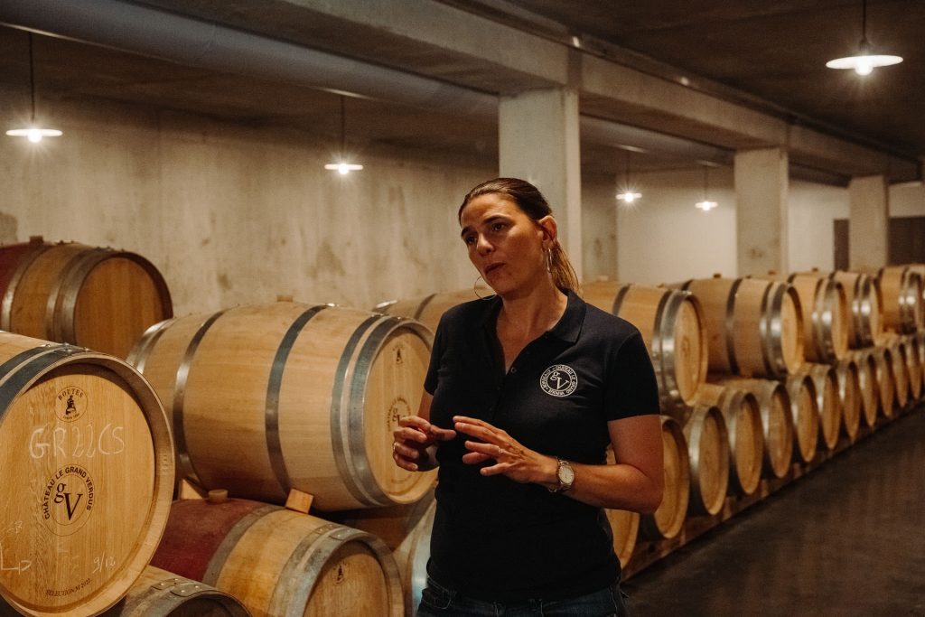 A woman in a navy polo shirt gestures while discussing Bordeaux wine, surrounded by stacked wooden barrels in a warmly lit wine cellar.