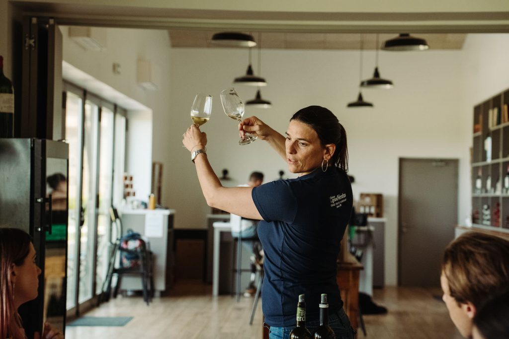 A woman in a navy shirt evaluates two glasses of red wine, addressing seated guests in a bright Bordeaux winery tasting room.
