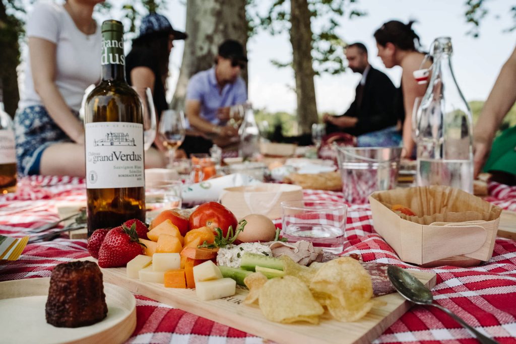 Several friends share Bordeaux wine, cheese, fruit, and snacks on a red-checkered blanket in a sunny city park.
