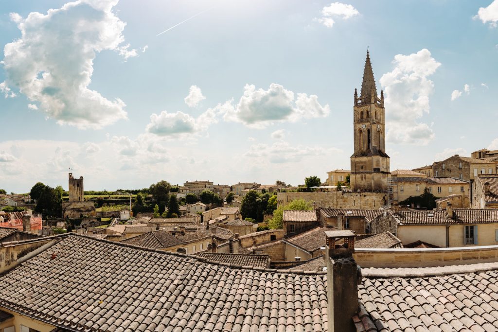 A historic European town features red-tiled rooftops, a tall stone church spire, and sunny skies over Saint-Émilion, France.