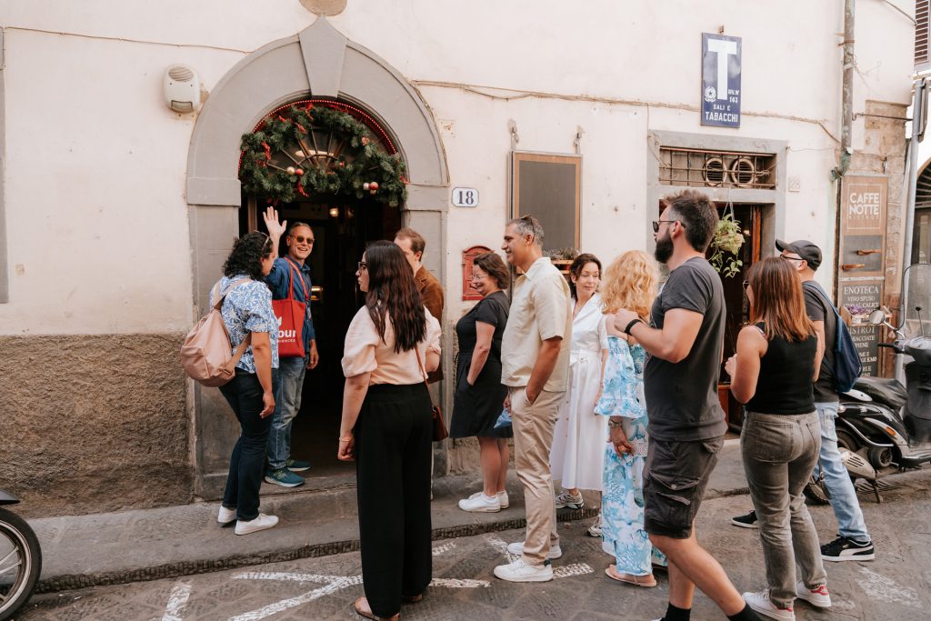 People wait in line outside a doorway decorated with a wreath on a narrow Tuscan street, chatting and looking inside.