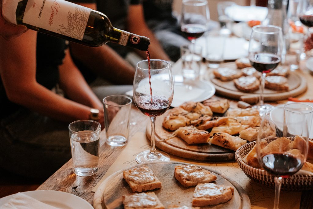 A hand pours red wine into a glass on a Tuscan table surrounded by breads, pastries, and friends enjoying a meal.