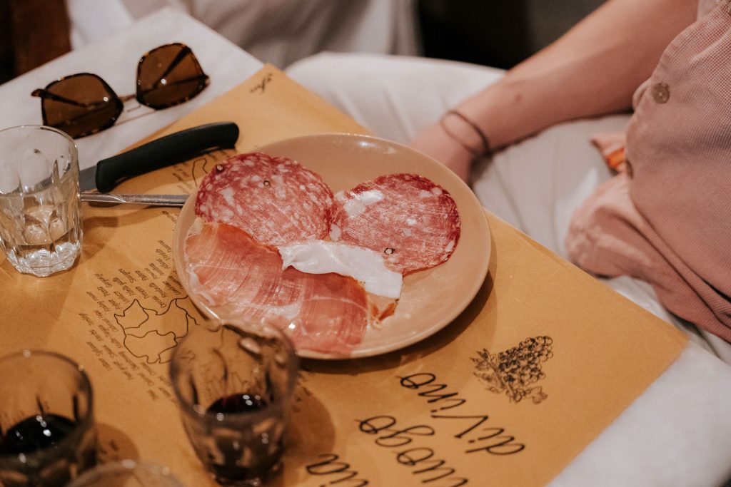 Several slices of cured meats and prosciutto rest on a plate atop brown paper, with empty glasses, cutlery, and sunglasses nearby.