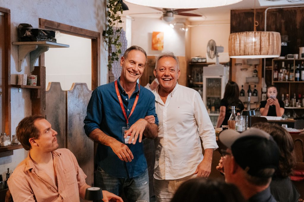 Two men smiling together in a warmly lit Tuscan restaurant, surrounded by guests engaged in lively conversation; inviting atmosphere.