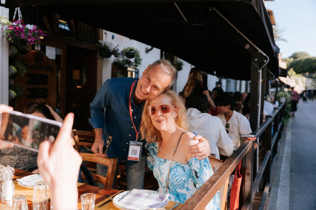 A man in a blue shirt and a woman in a blue dress with pink sunglasses smile together at an outdoor Tuscan restaurant.