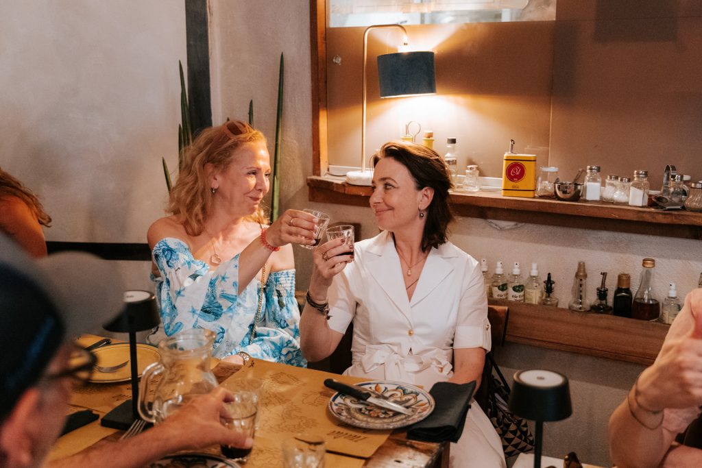 Two women toast at a Tuscan restaurant table, smiling as they enjoy drinks; shelves behind them hold bottles and jars.