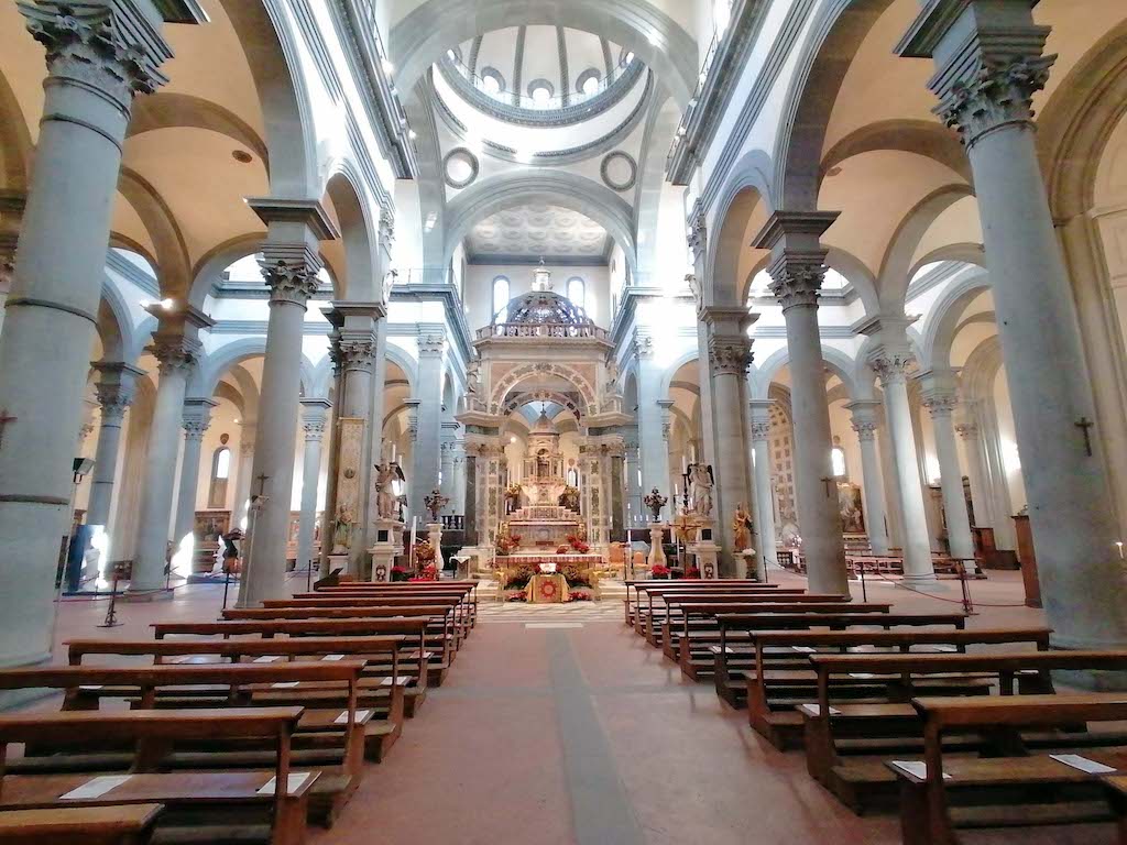 A grand church in Tuscany features high arched ceilings, stone columns, wooden pews, and an ornate altar with religious statues.