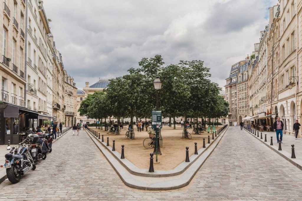 A peaceful triangular plaza in Paris features trees, benches, and historic buildings; people stroll nearby as motorcycles are parked.