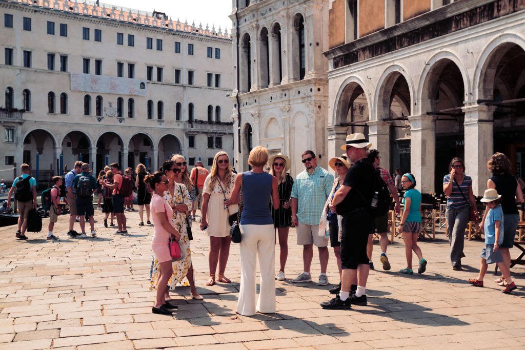 A tour guide explains local history to tourists in a sunny European square lined with arched, historic buildings and bustling visitors.