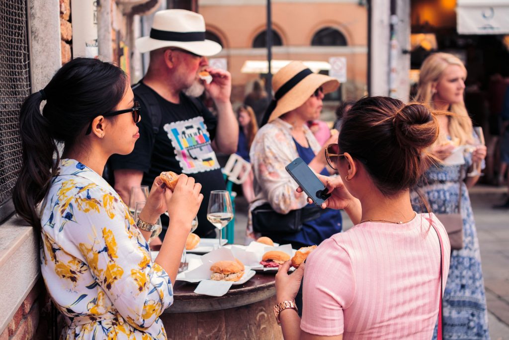 Several people sit at an outdoor cafe table in a sunny city. Two women in floral dresses talk and check a smartphone.