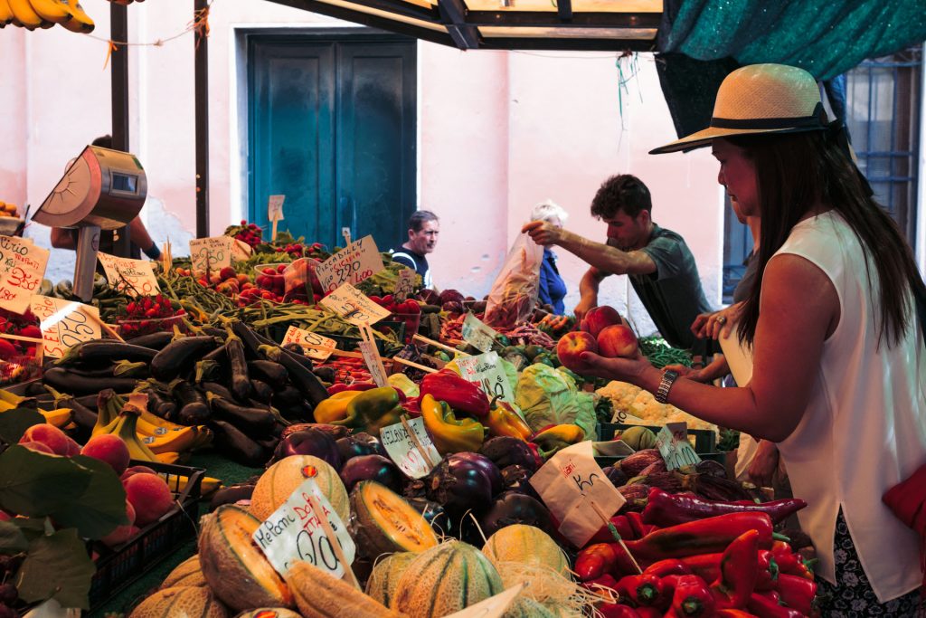 A woman in a straw hat picks fruit at a bustling outdoor market stand in Barcelona, surrounded by colorful produce and handwritten price signs.
