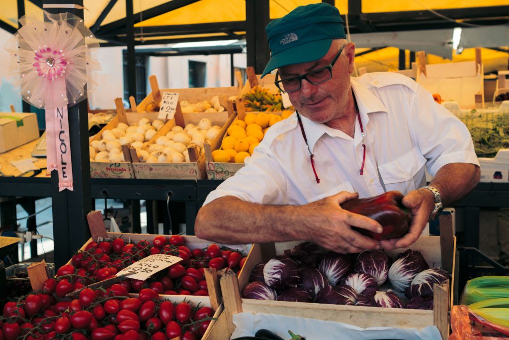 An older man in a teal cap chooses vegetables at an outdoor market stall labeled “NICOLE,” surrounded by fresh produce in Venice.