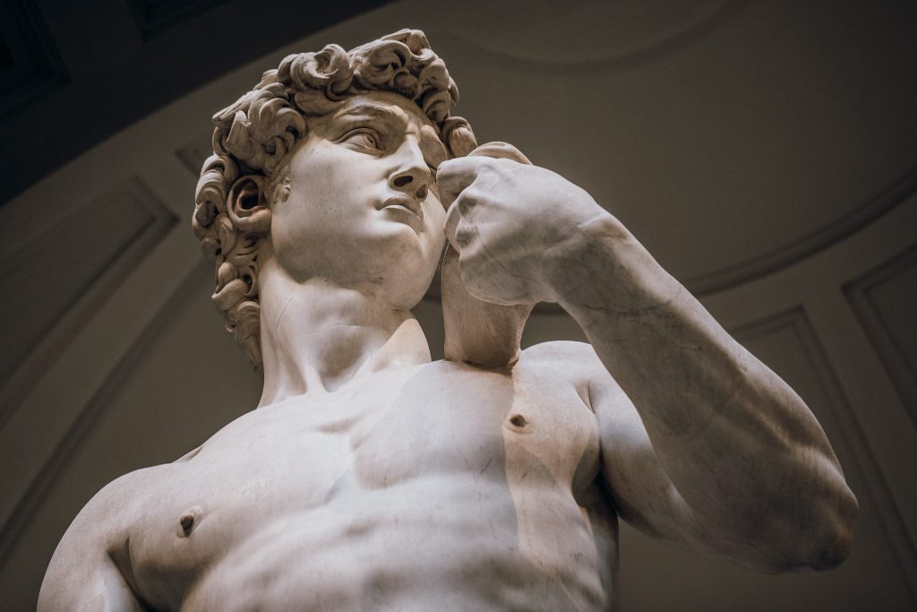 A marble statue of David by Michelangelo stands in Florence, showing his muscular torso, curly hair, and tense right arm.