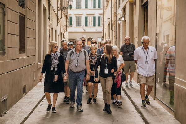 Tourists walk down a narrow street in Florence, wearing casual clothes and audio guides, surrounded by historic buildings with shutters.