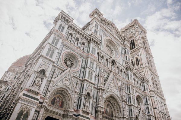 The Florence Cathedral’s colorful marble facade displays intricate carvings, statues, arches, and rose windows under a partly cloudy sky.