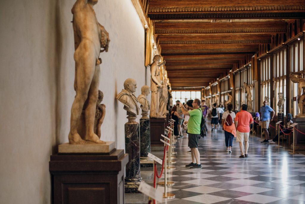 Visitors explore Florence’s Uffizi Gallery, admiring marble statues and busts in a grand hall with checkered floor and wooden ceiling.