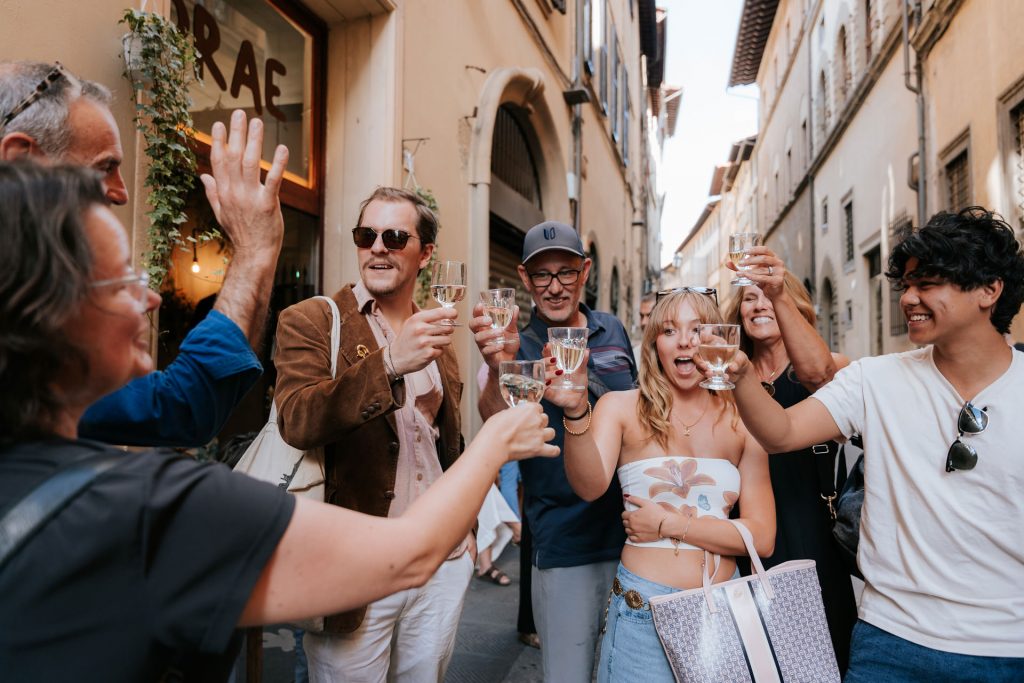 Five friends toast with wine glasses on a sunny, narrow street in Florence, Italy, celebrating together and smiling warmly.