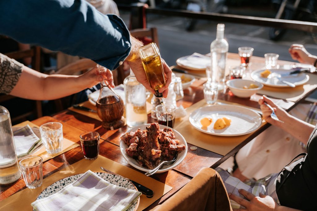 At a sunny Tuscan table, someone pours olive oil onto grilled meat as friends gather and share a lively outdoor meal.