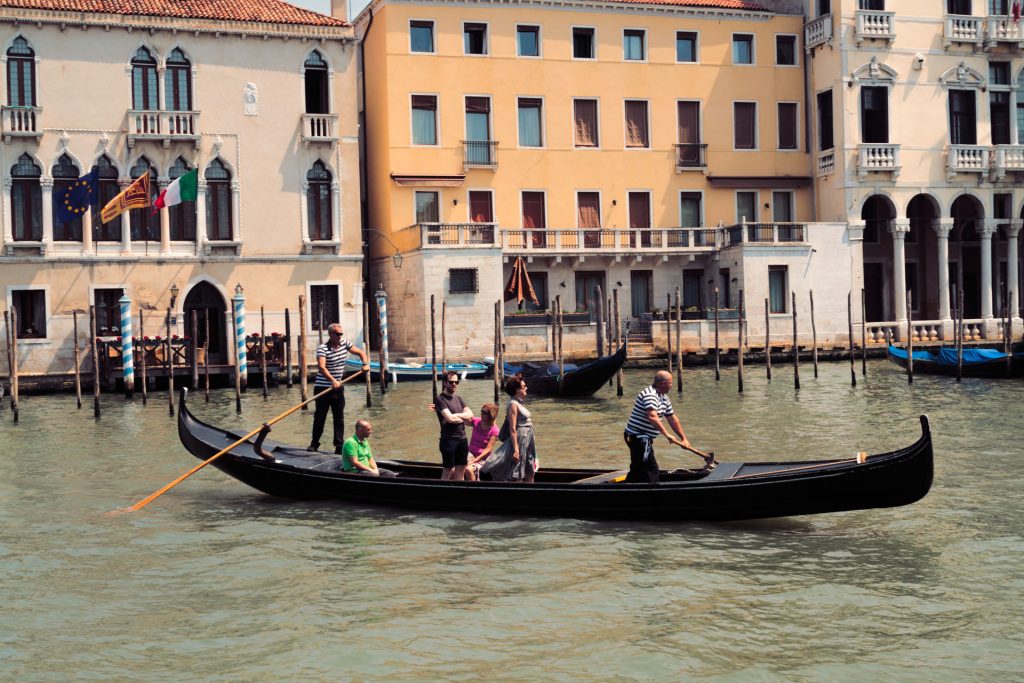 A gondolier steers a gondola carrying five people through a Venice canal, passing historic buildings and boats along the water.